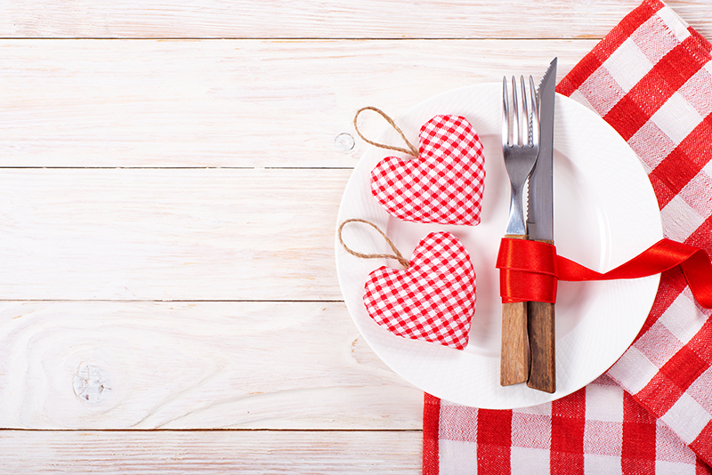 Plato blanco con cubiertos y adornos de corazones. Debajo, un mantel con cuadros rojos y blancos sobre madera.