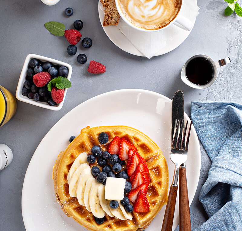 Plato de waffles con arándanos, fresas, banano y mantequilla, acompañado con una taza de café cremoso. En la vista, un panal de huevos, cubiertos y syrup.
