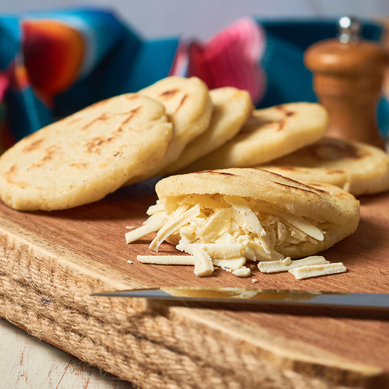 Arepas de maíz, rellenas de queso rallado sobre una bandeja de madera. Al frente hay un cuchillo y al fondo se ve una manta de colores.