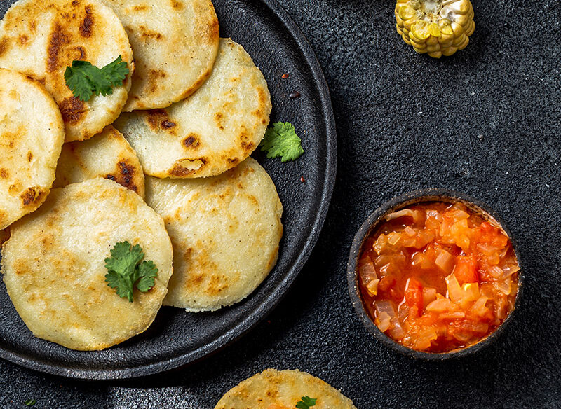Arepas de maíz de trigo sobre un plato negro con especias, acompañadas de una arepa con tomate picado, mazorcas de maíz. Todo sobre un fondo oscuro.