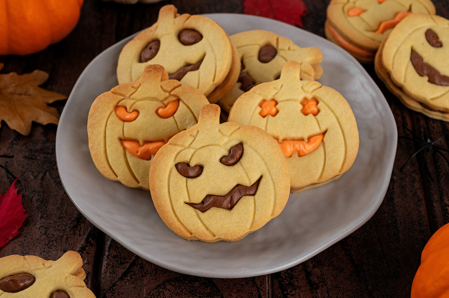 Galletas de maíz horneadas, rellenas con chocolate y crema pastelera sobre un plato gris. Debajo del plato, se encuentra una mesa de madera oscura, más galletas, hojas secas de árbol y una calabaza.